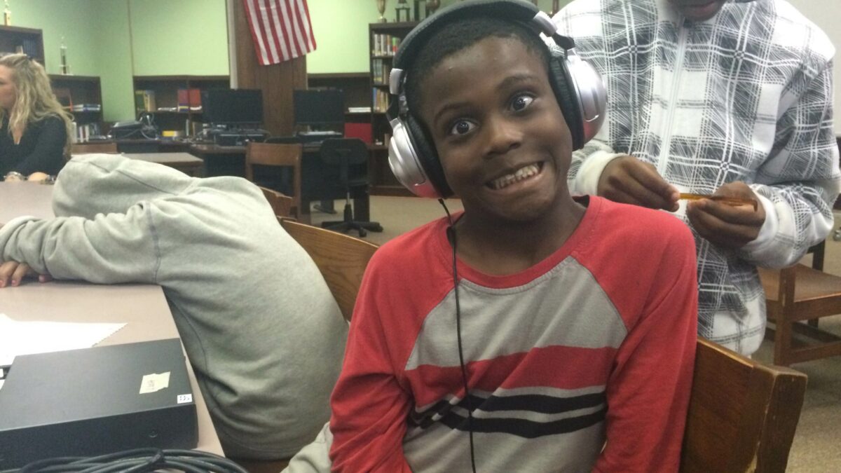 A youth making a silly face wearing a pair of headphones sitting on a wooden chair in a library