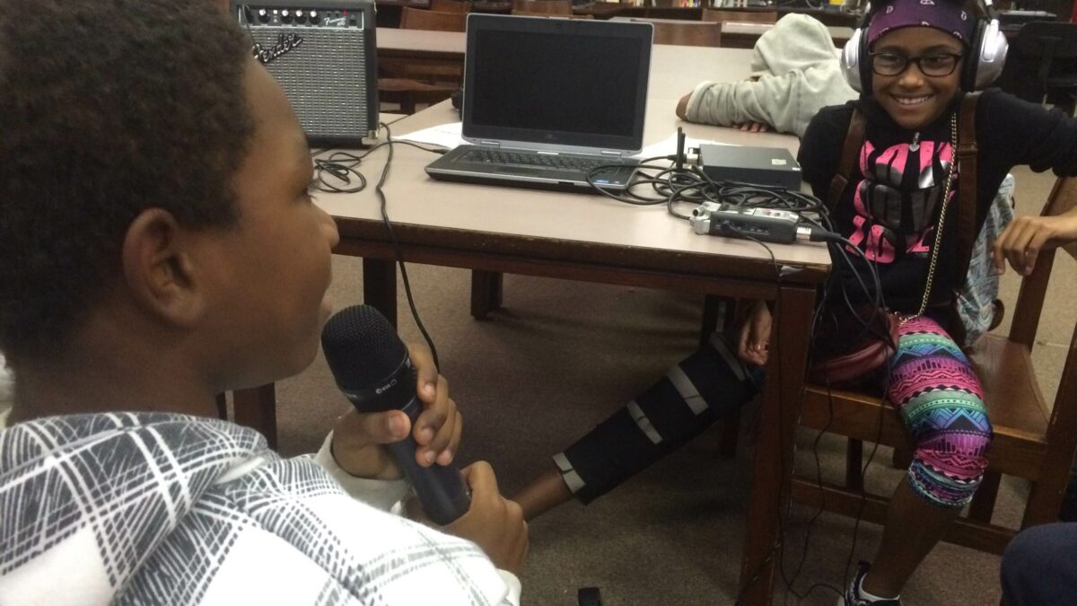 A youth holding and speaking into a microphone and another youth wearing headphones watching in a library