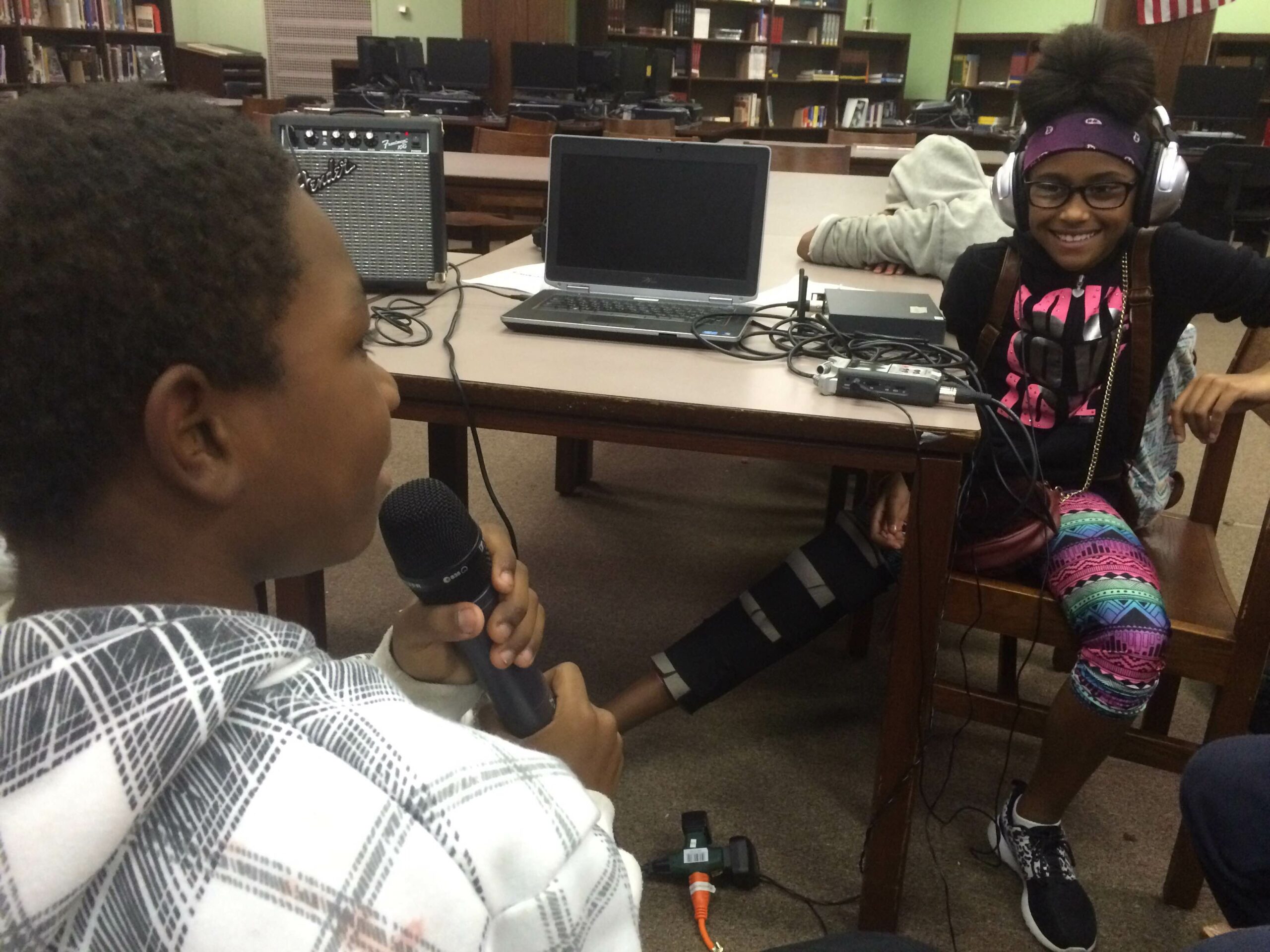 A youth holding and speaking into a microphone and another youth wearing headphones watching in a library