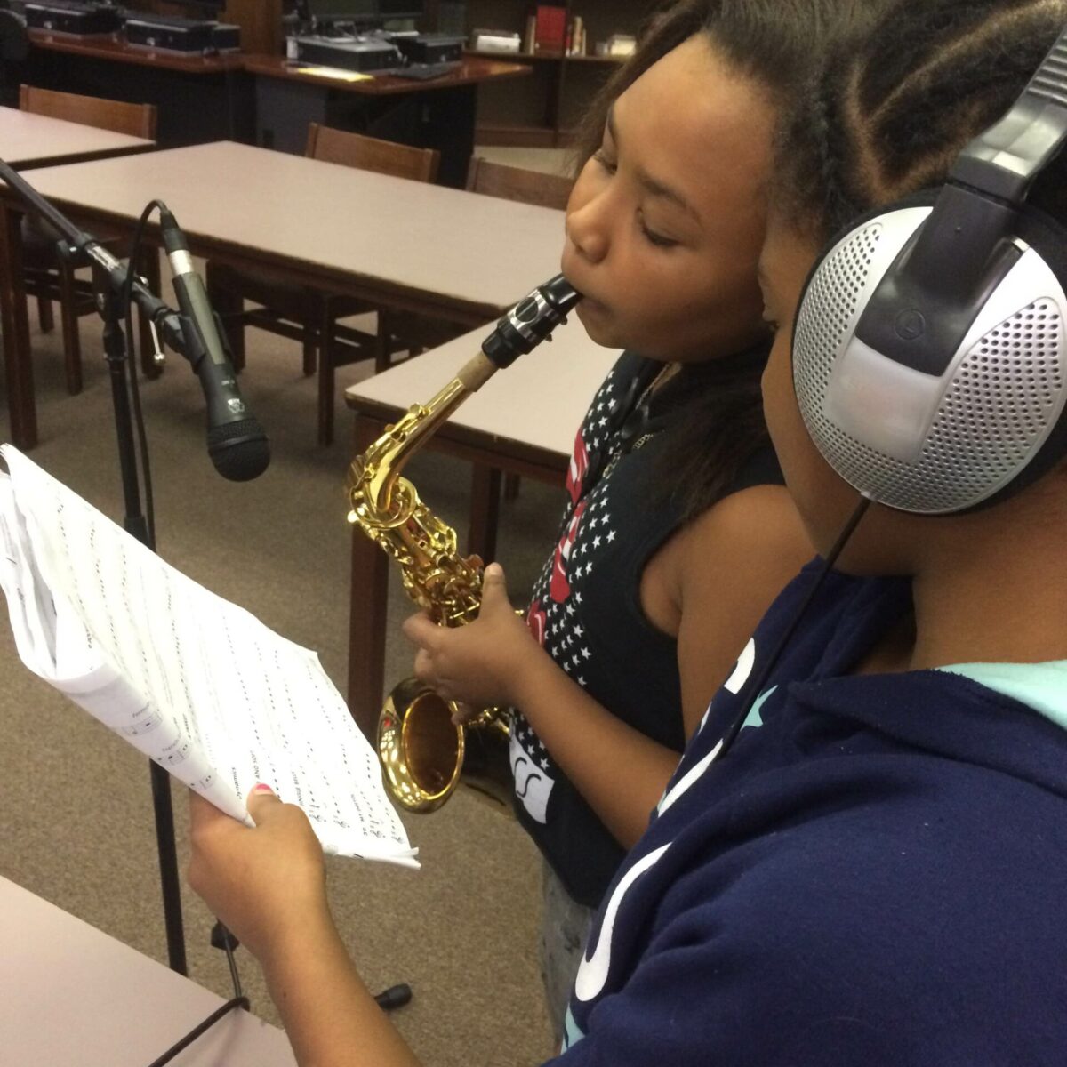 IMG_1918 A youth wearing headphones holding a piece of paper looking at another youth playing an alto saxophone in a microphone in a classroom
