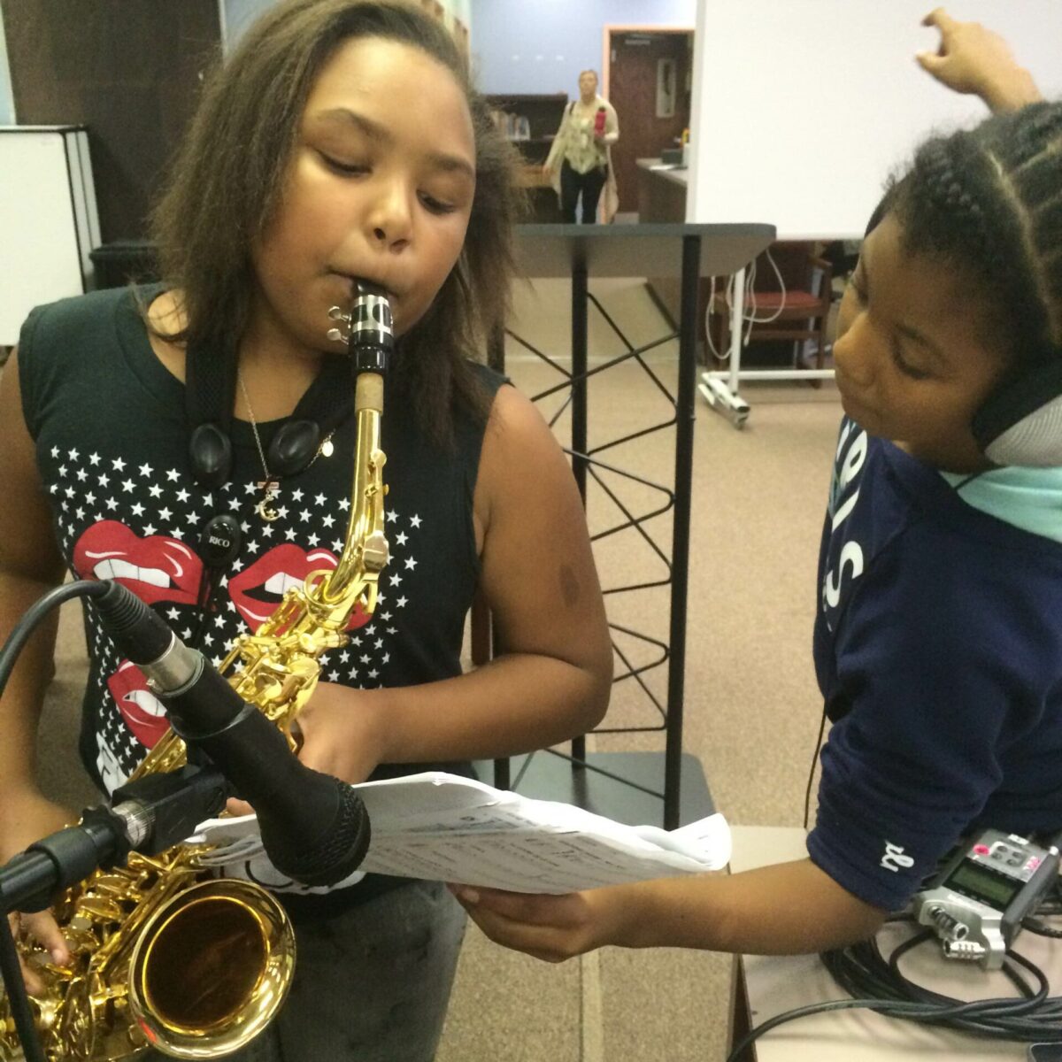 IMG_1921 A youth wearing headphones holding a piece of paper looking at another youth playing an alto saxophone in a microphone in a classroom