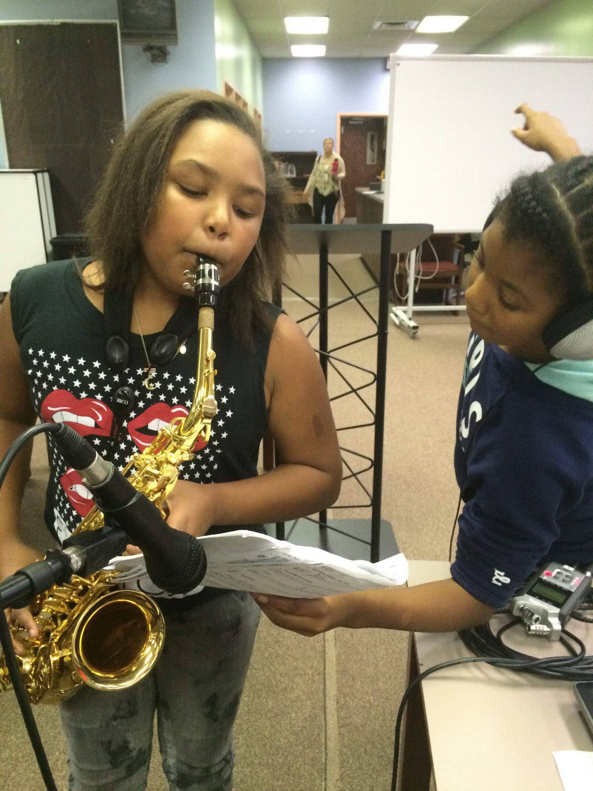A youth wearing headphones holding a piece of paper looking at another youth playing an alto saxophone in a microphone in a classroom