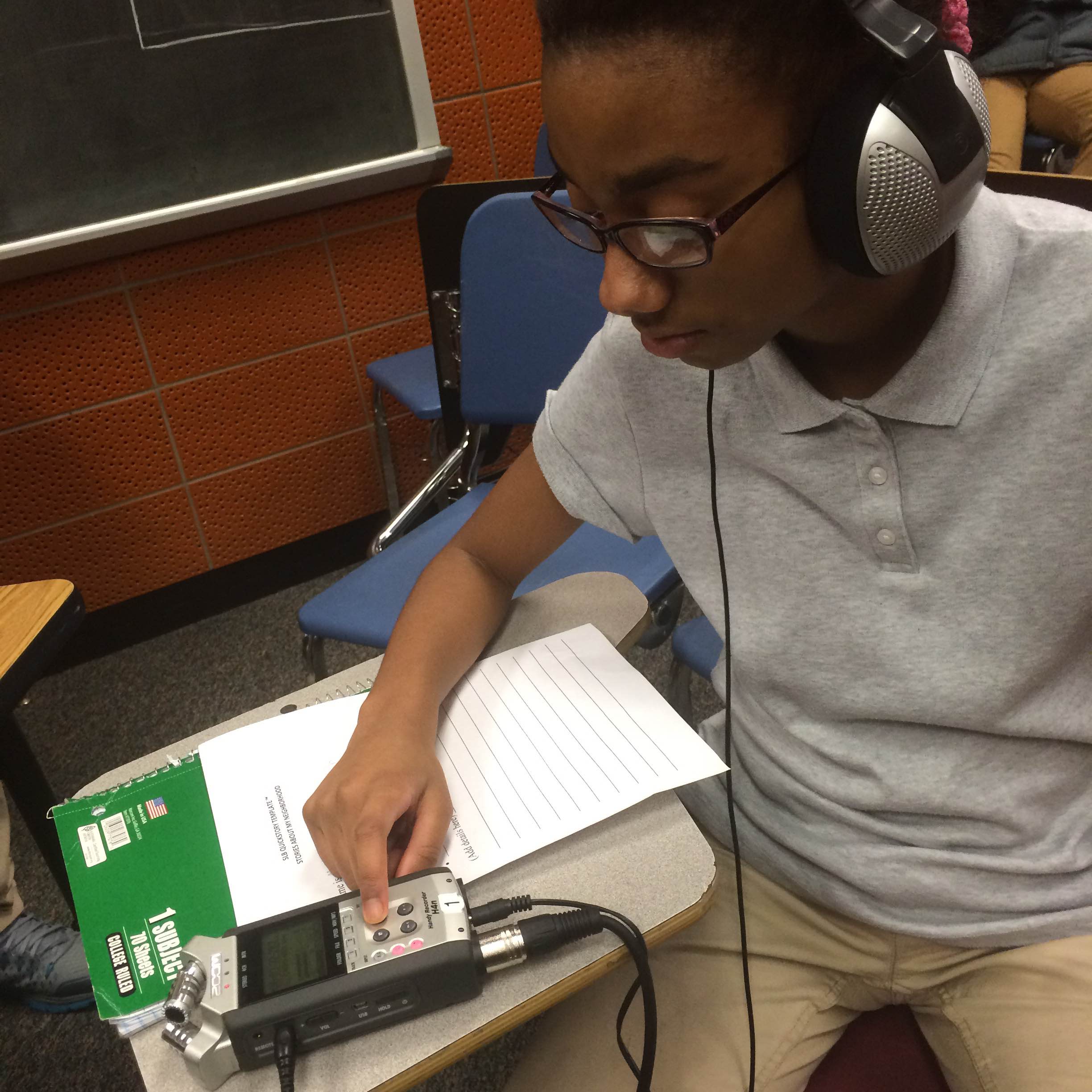 A youth wearing headphones interacting with a portable recorder looking down at a piece of paper on a classroom desk