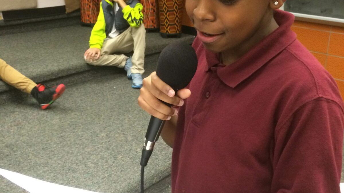 A youth holding and speaking into a microphone and holding a piece of paper in a classroom