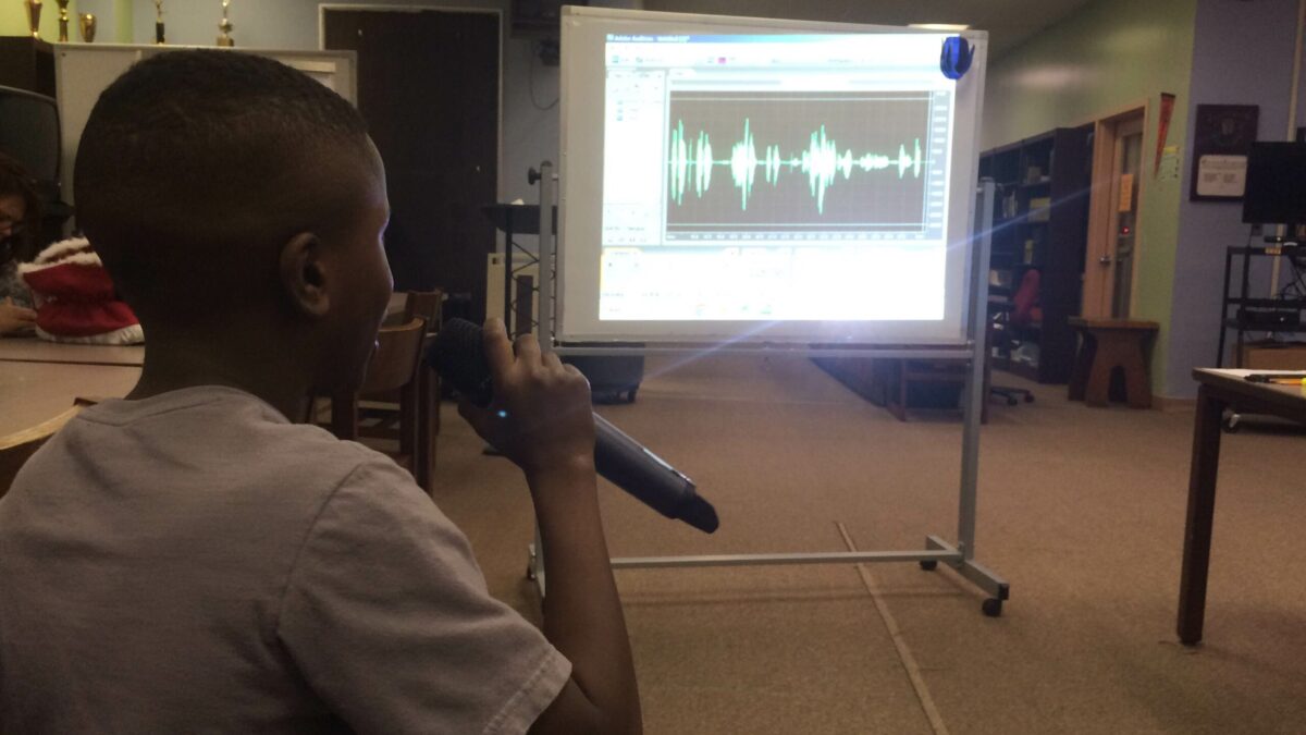 IMG_2024 A youth holding and speaking into a microphone looking at audio editing software on a projected screen in a library