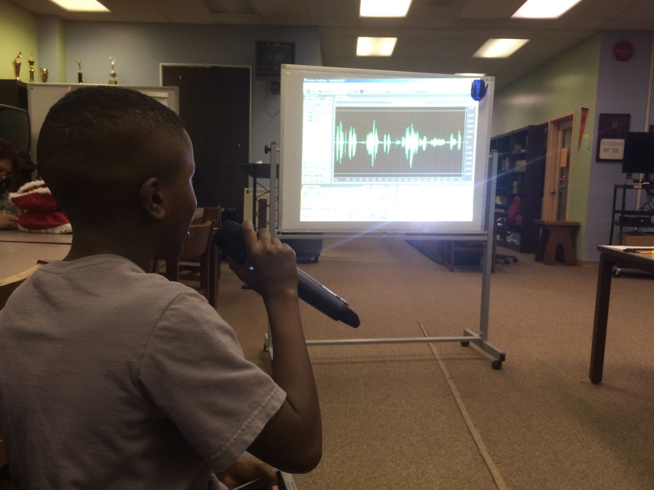 A youth holding and speaking into a microphone looking at audio editing software on a projected screen in a library