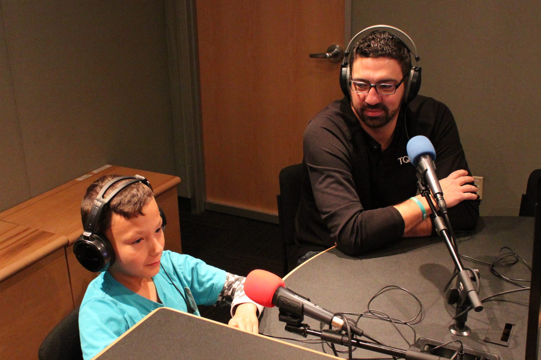 A youth and an adult wearing headphones speaking into microphones in a radio studio