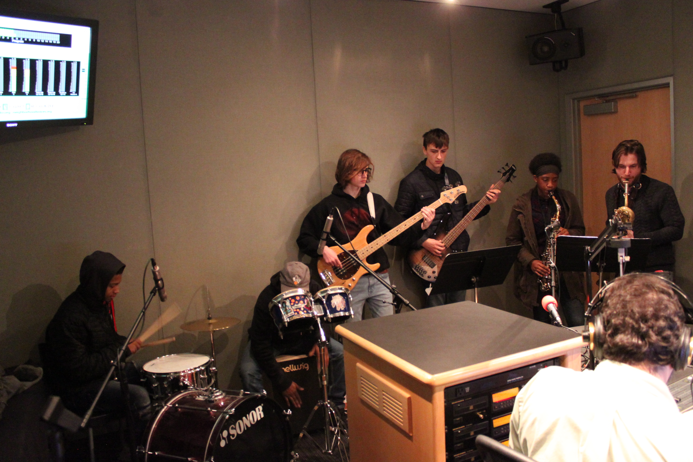 A teen playing drum set, a teen playing a cajon, a teen playing electric bass, a teen playing electric guitar, a teen playing alto saxophone, and an adult playing baritone saxophone as an adult wearing headphones listens in a radio studio