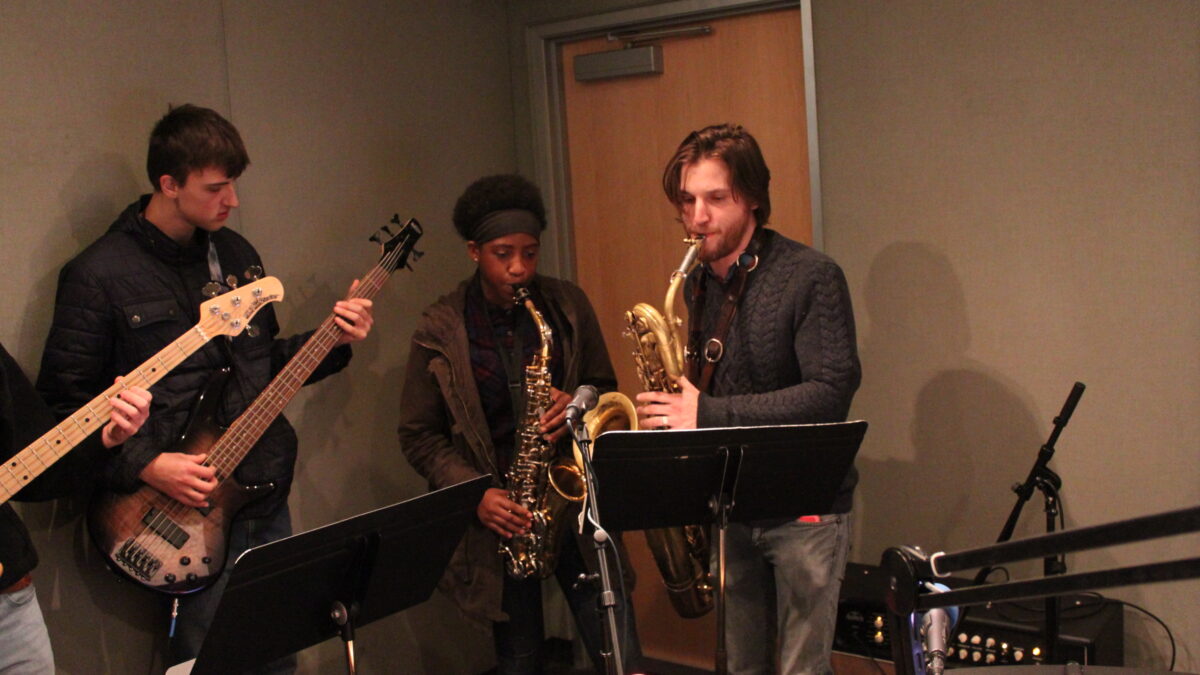 A teen playing electric guitar, a teen playing alto saxophone, and an adult playing baritone saxophone in a radio studio