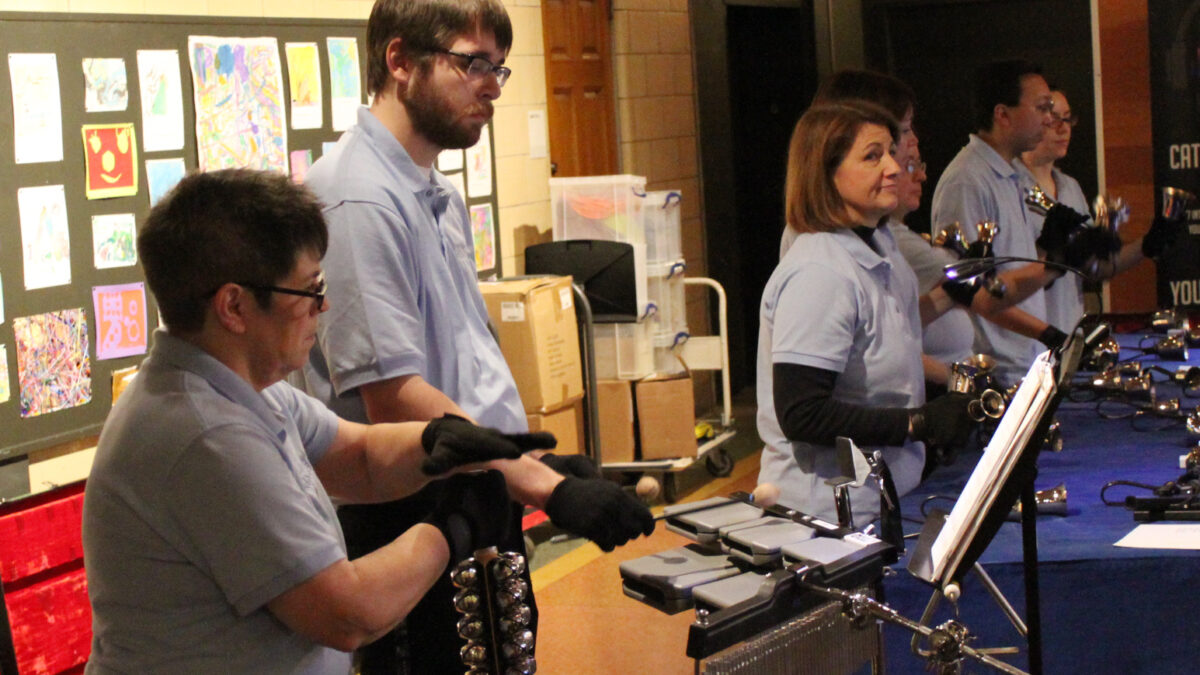 An adult playing jingle bells, an adult playing cowbells, and five adults playing handheld bells