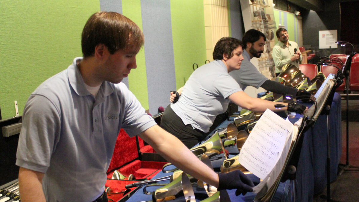 Three adults standing in front of bells flipping pages on music stands