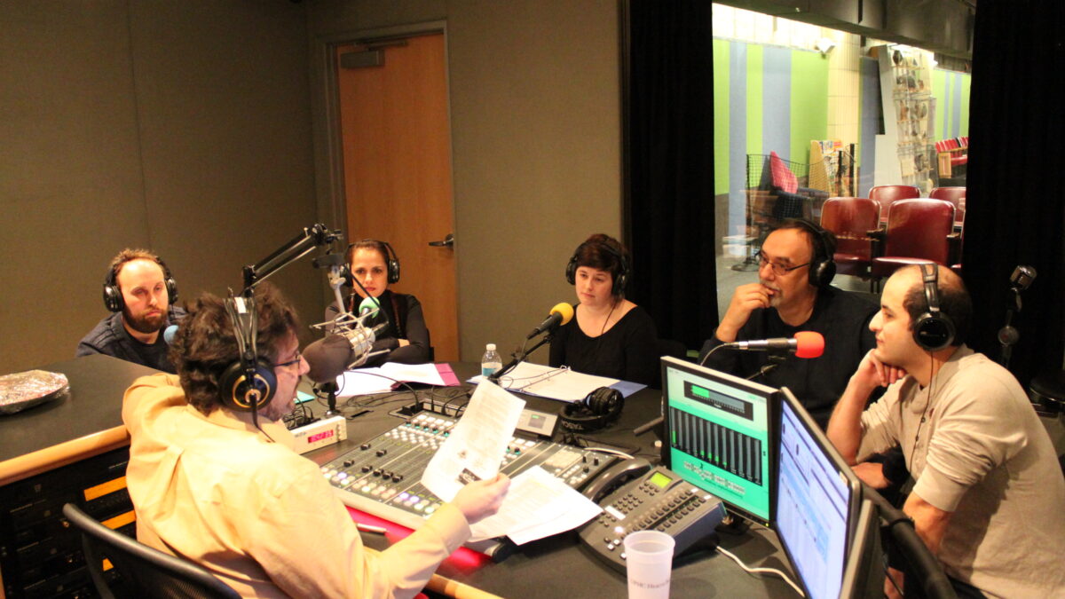 Five adults wearing headphones sitting behind microphones being interviewed by an adult wearing headphones sitting behind a microphone and a mixing board in a radio studio