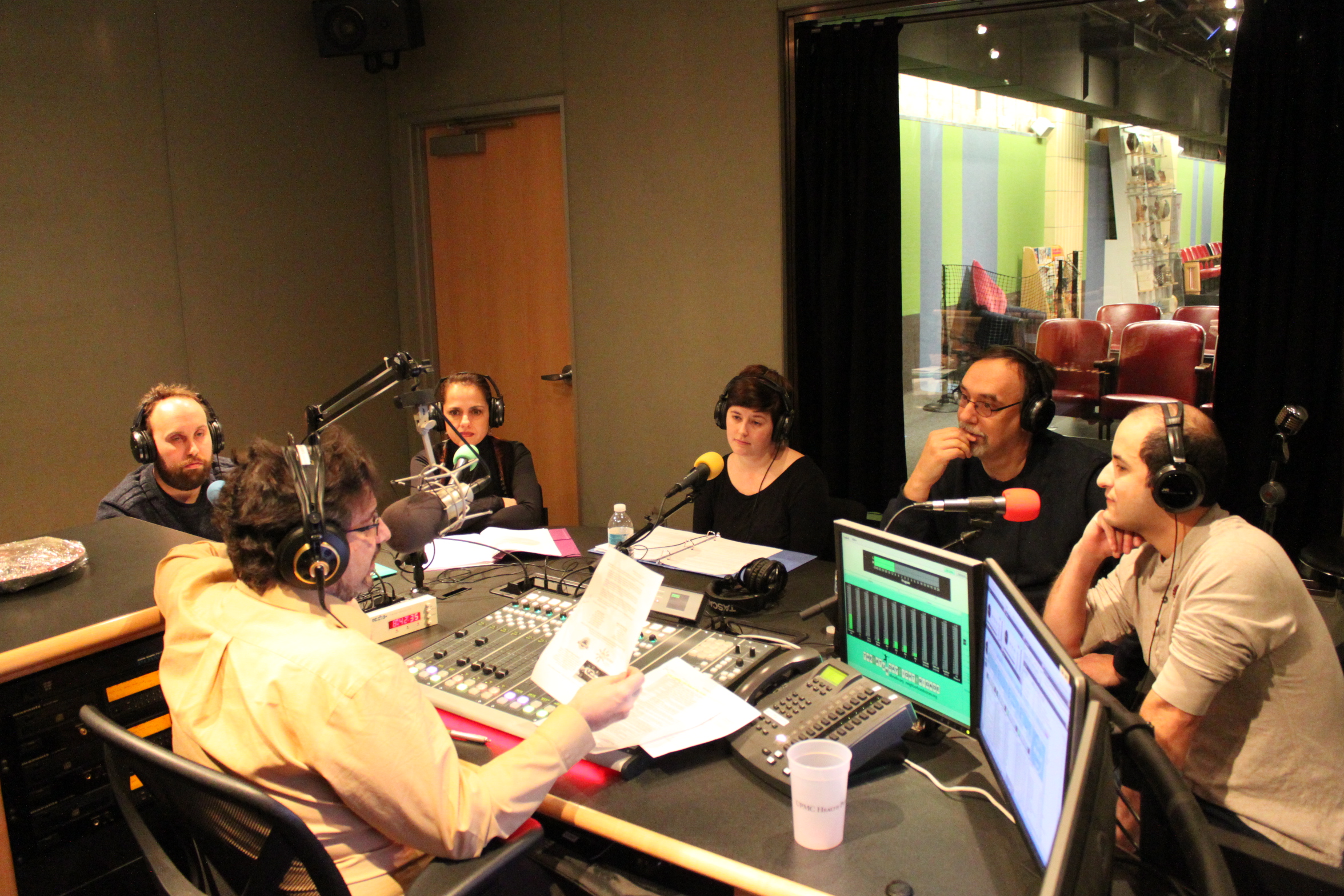 Five adults wearing headphones sitting behind microphones being interviewed by an adult wearing headphones sitting behind a microphone and a mixing board in a radio studio