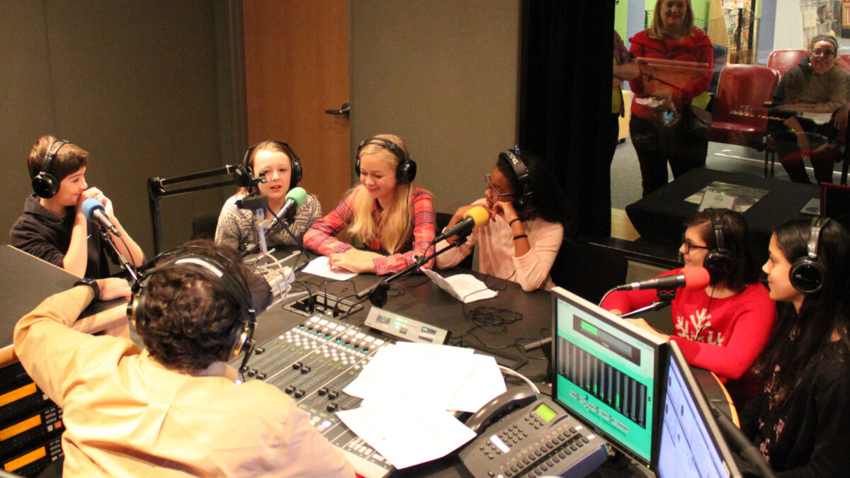 IMG_5114 Six youth wearing headphones speaking into microphones being interviewed by an adult wearing headphones sitting behind a microphone and a mixing board in a radio studio