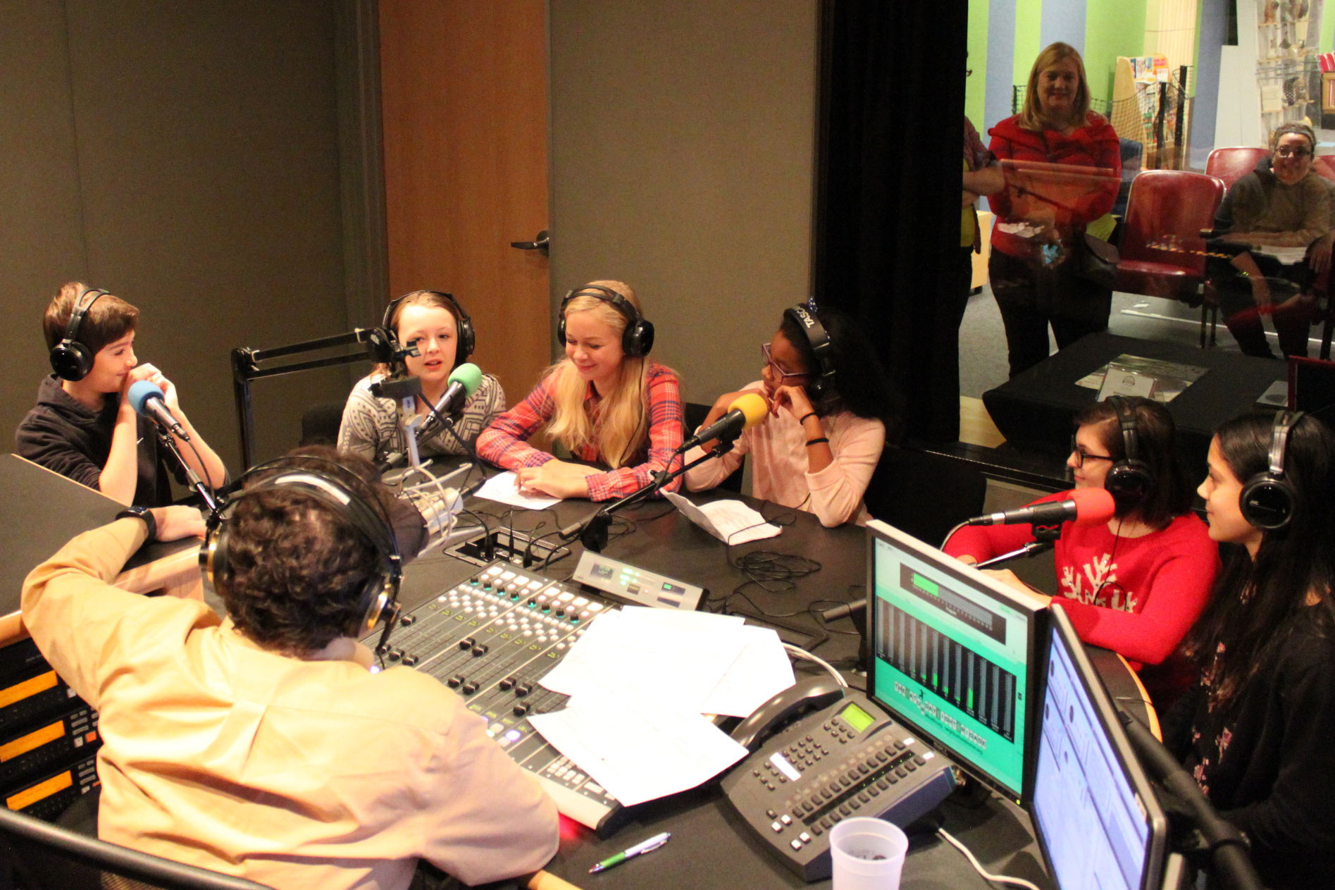 Six youth wearing headphones speaking into microphones being interviewed by an adult wearing headphones sitting behind a microphone and a mixing board in a radio studio