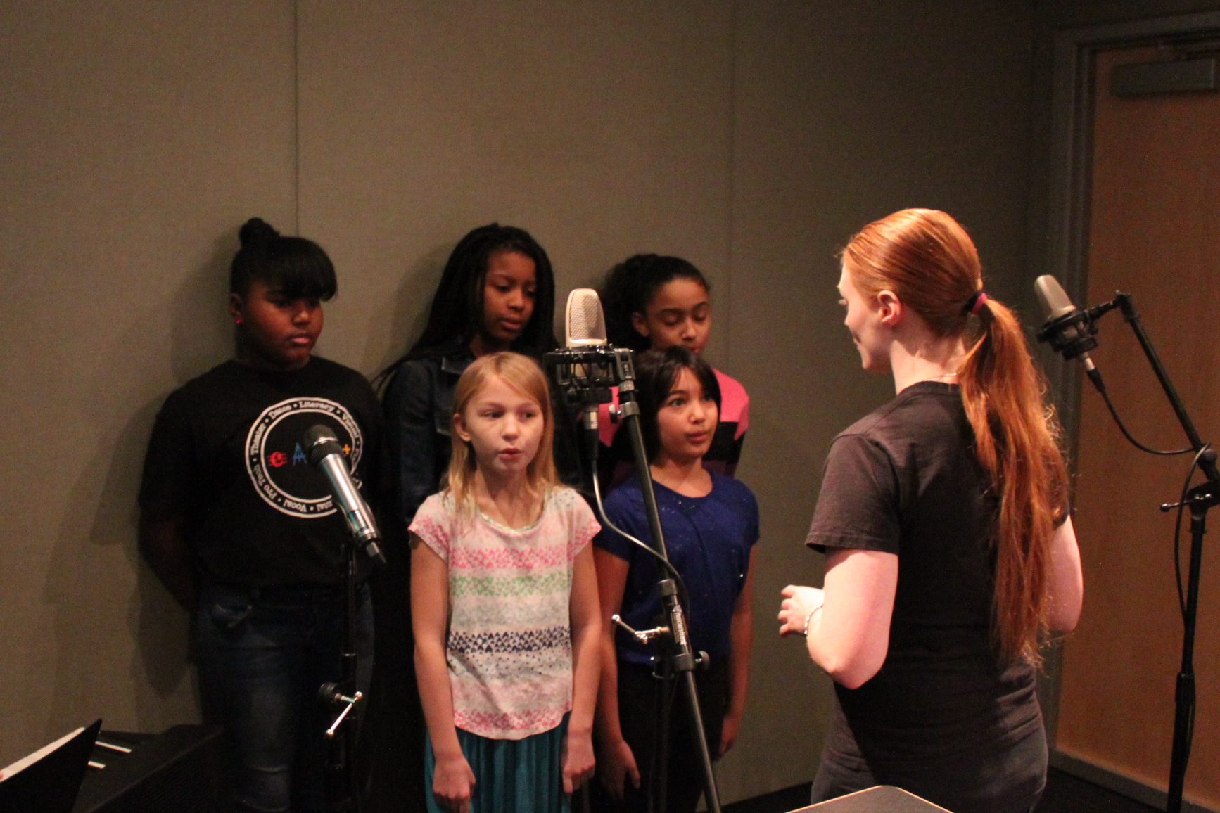 Five youth singing into a microphone being led by an adult in a radio studio