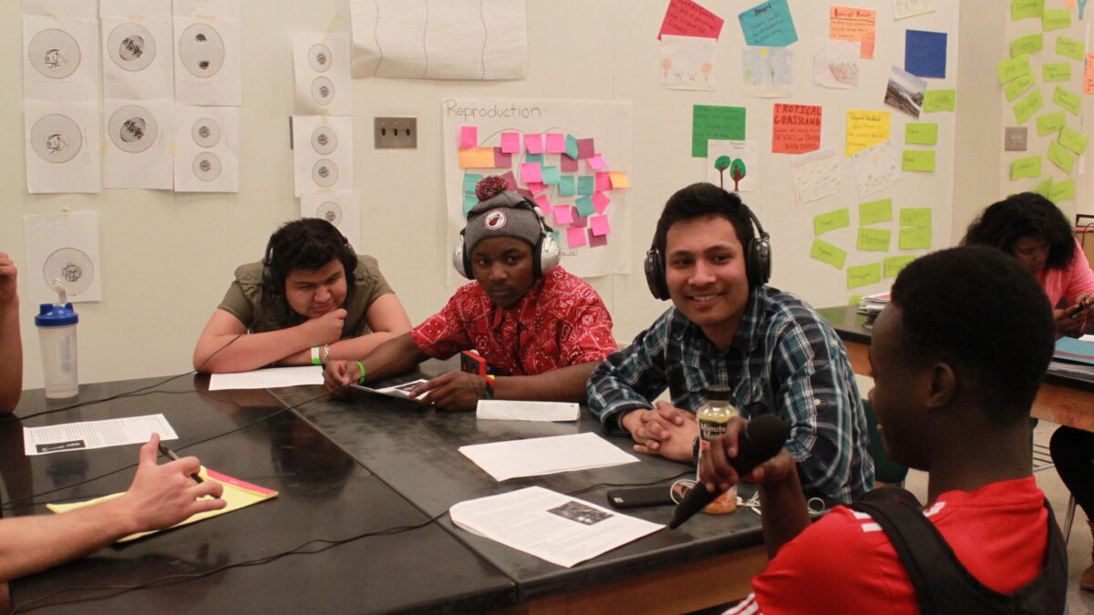 IMG_7814 Four teens sitting around pieces of paper on a classroom desk, one holding and speaking into a microphone