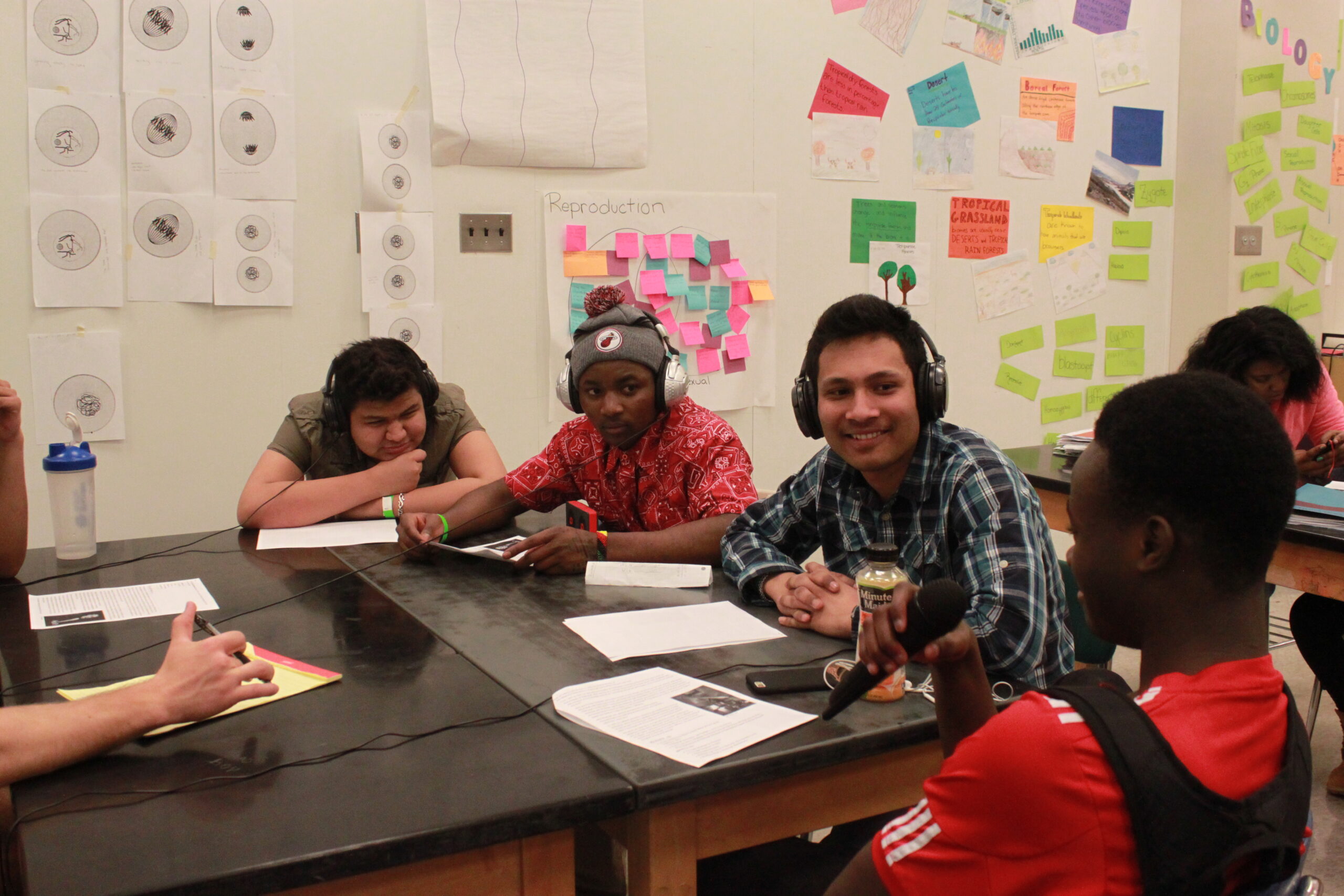 Four teens sitting around pieces of paper on a classroom desk, one holding and speaking into a microphone