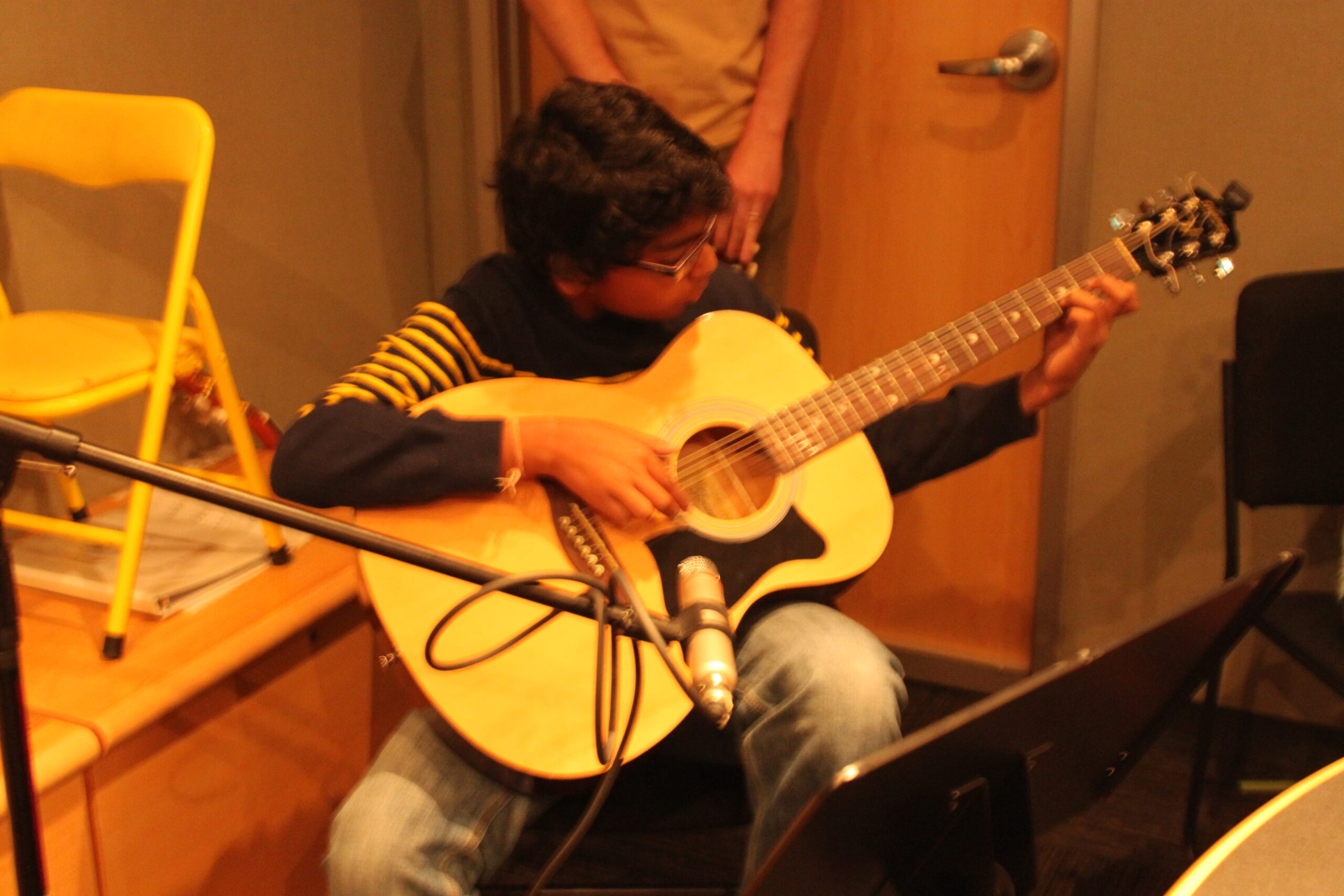A youth playing an acoustic guitar sitting behind a music stand in a radio studio