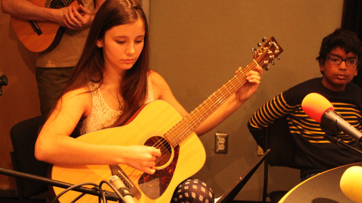 IMG_8810 A youth playing an acoustic guitar looking at a music stand as another youth sitting in the background watches in a radio studio