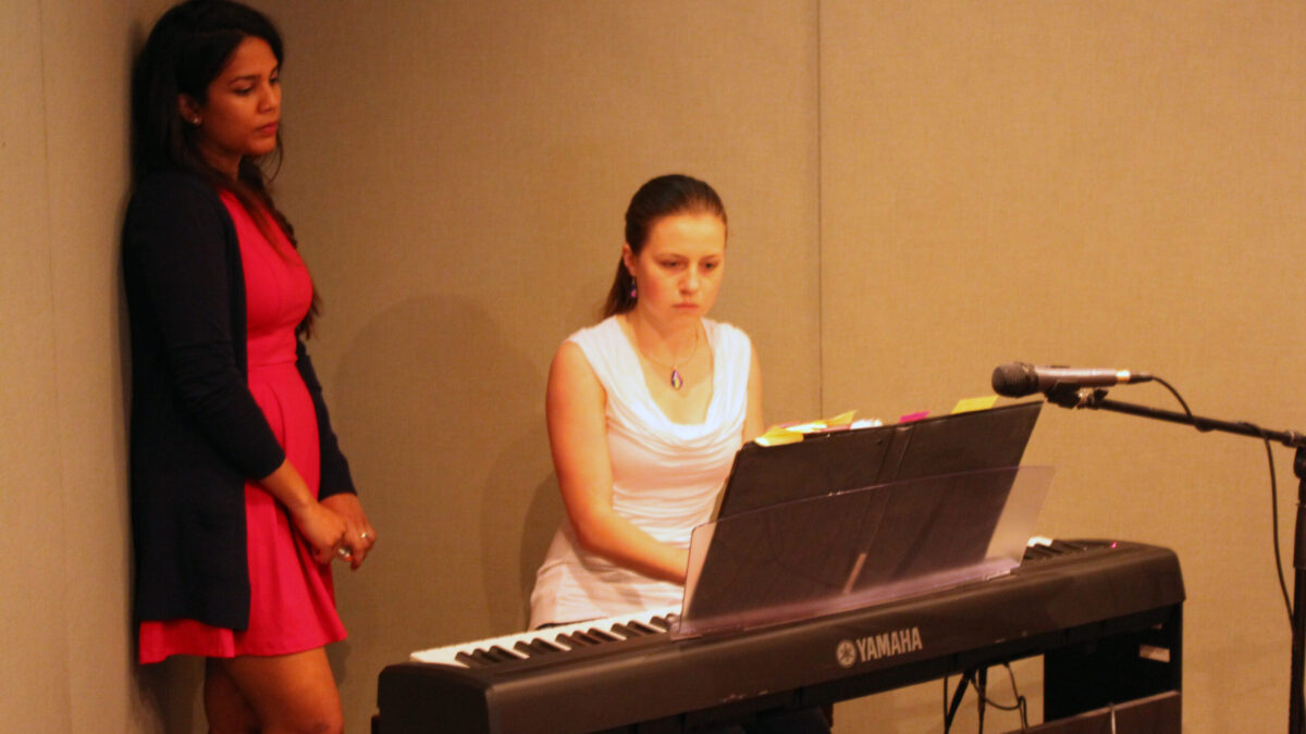 An adult looking at another youth playing a piano keyboard sitting behind a microphone in a radio studio