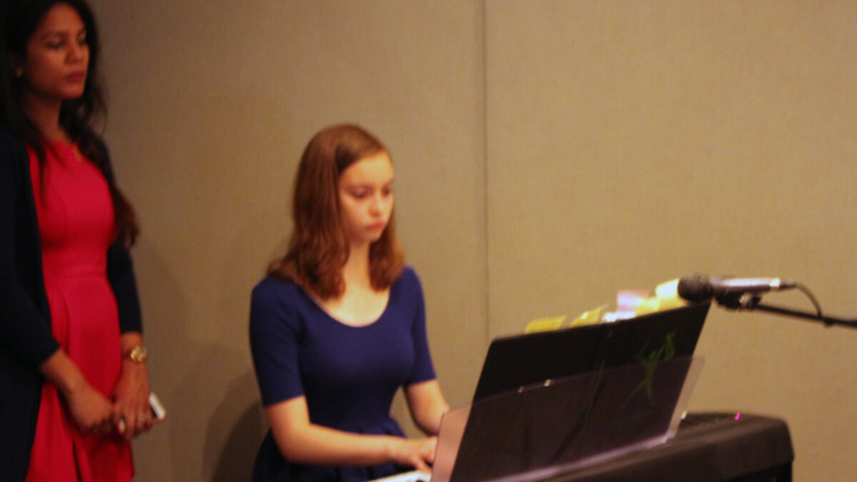 An adult watching a youth playing a piano keyboard sitting behind a microphone in a radio studio