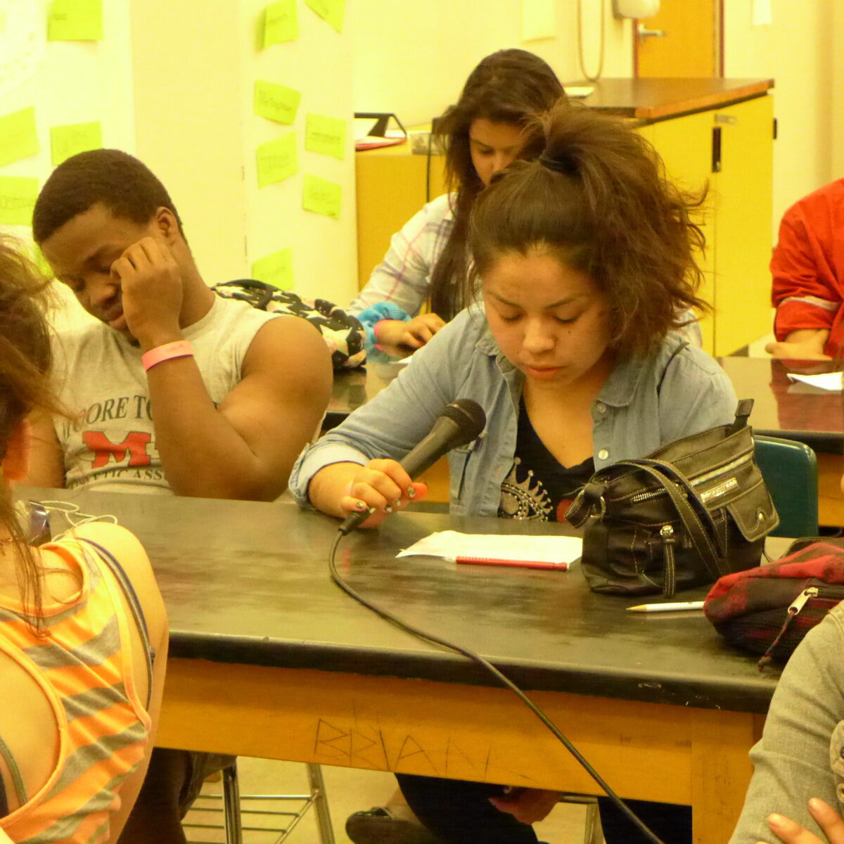Three youth sitting at classroom desks, one holding a microphone while looking down at a piece of paper