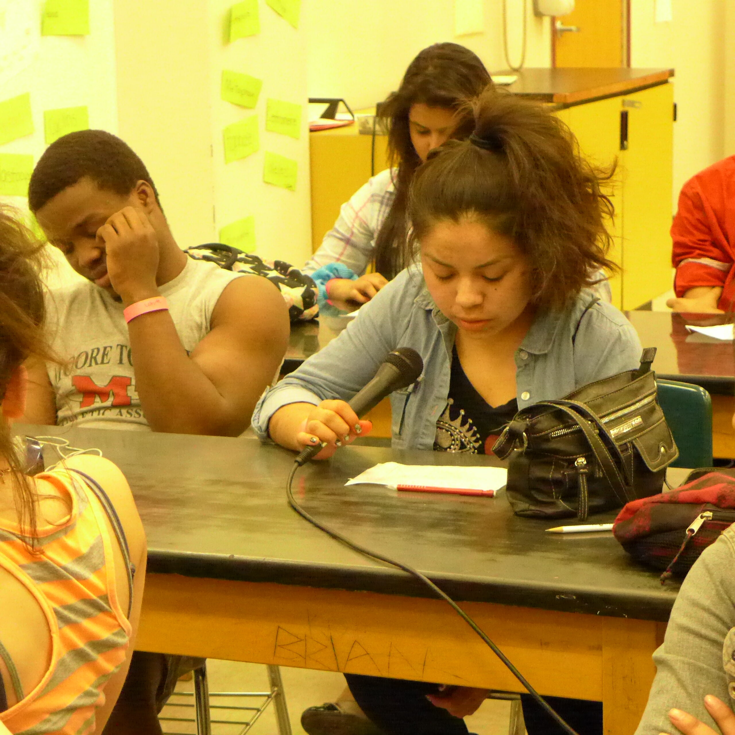 Three youth sitting at classroom desks, one holding a microphone while looking down at a piece of paper
