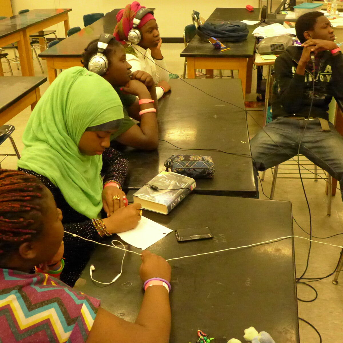 Four youth sitting at classroom desks, two wearing headphones, one looking down and writing on a piece of papeer