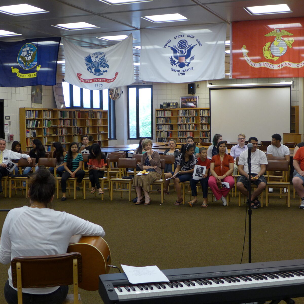 Fifteen individuals sitting in the audience listening to an individual playing an acoustic guitar in a library