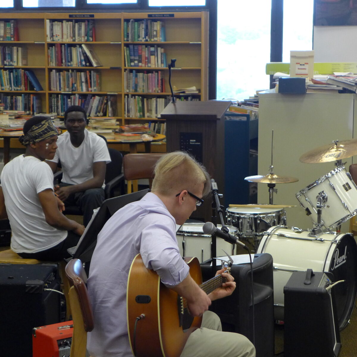 Two teens sitting in the background watching an individual playing an acoustic guitar in a library