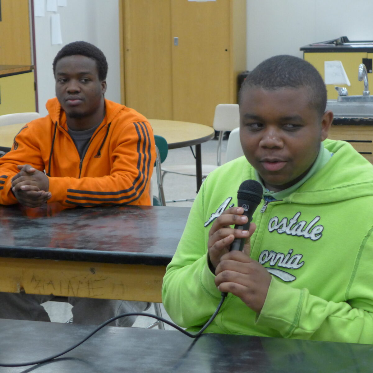 P1020638 Two teens sitting in a classroom, one holding and speaking into a microphone