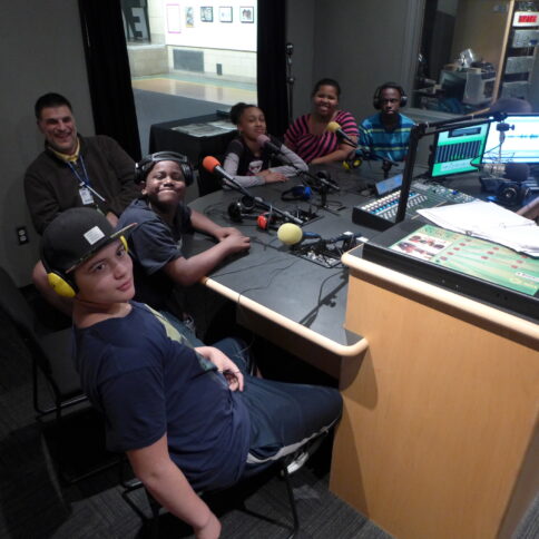 Five youth wearing headphones sitting behind microphones and one adult, all in a radio studio