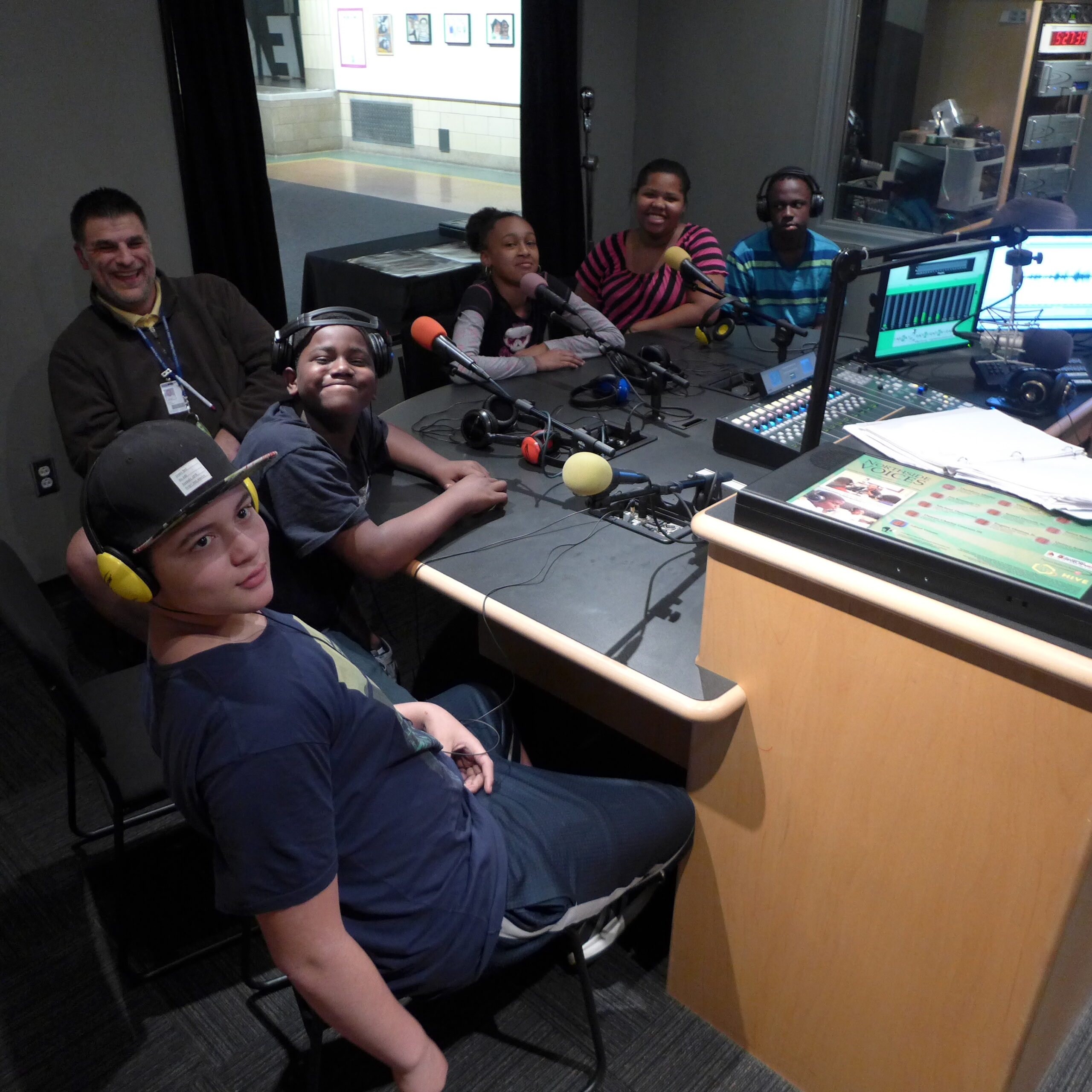 Five youth wearing headphones sitting behind microphones and one adult, all in a radio studio