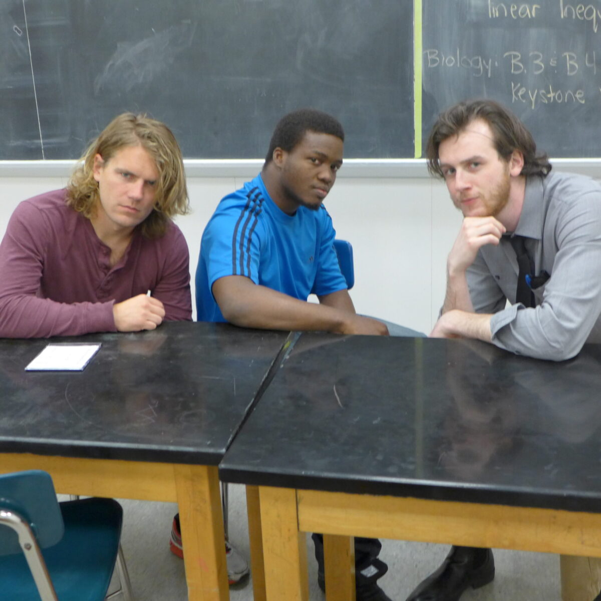 Two adults and a teen sitting and posing at a classroom desk in front of a chalkboard