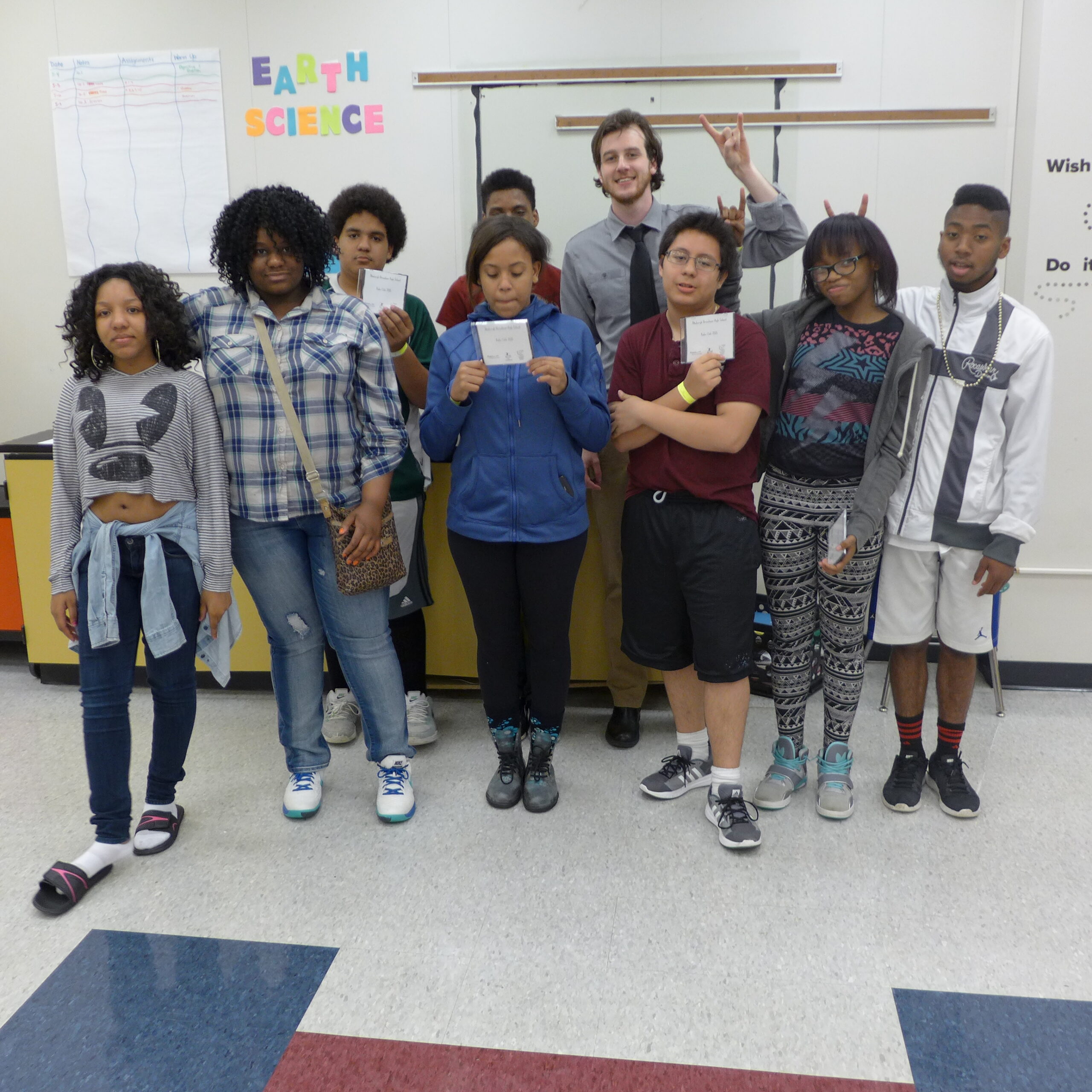 Eight teens, three teens holding cd cases, and one adult making a rocker hand sign, all standing in profile