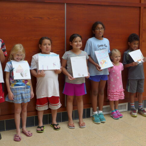 A group of youth standing in front of a wooden wall holding crayon drawings