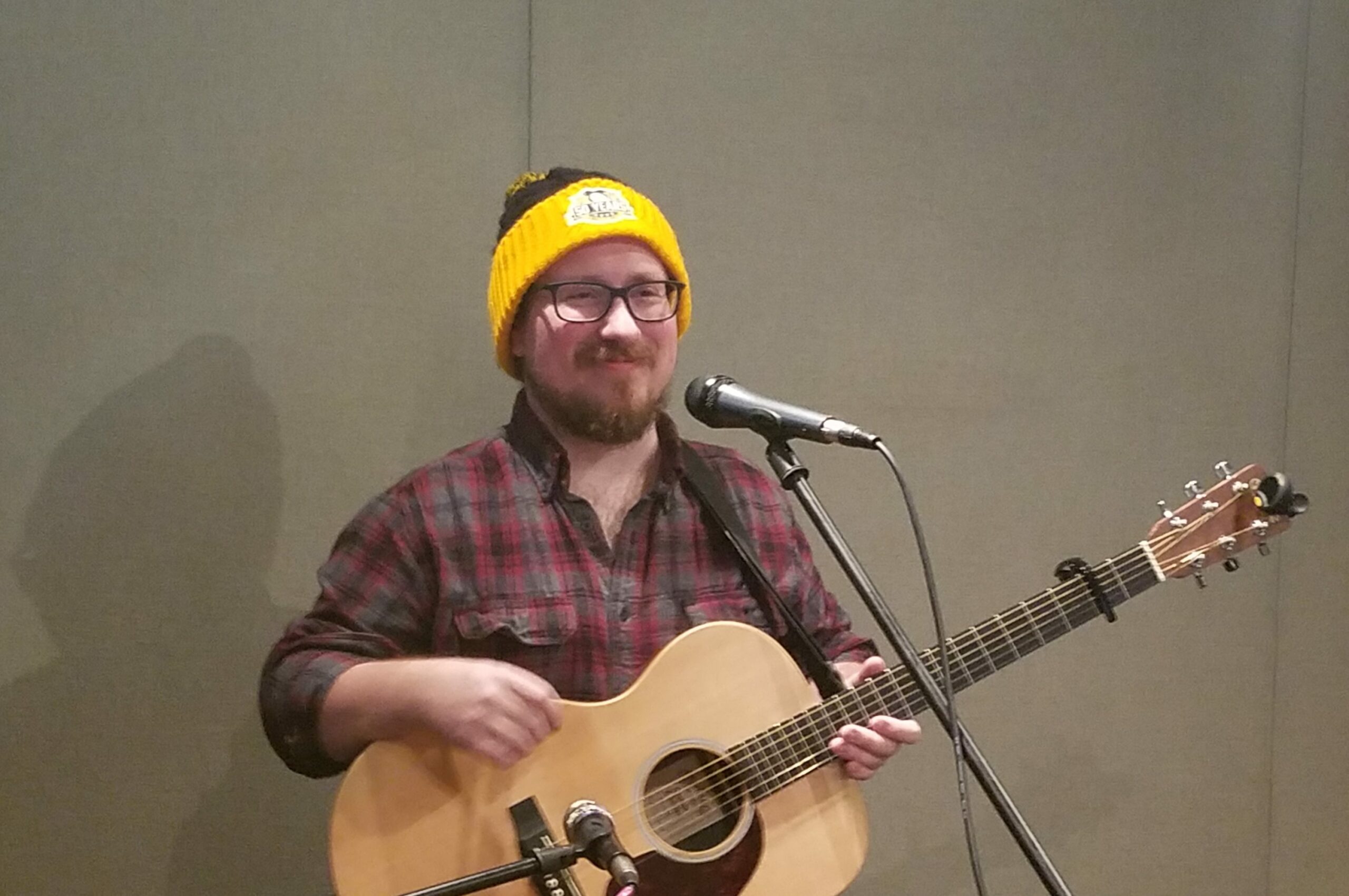 An adult playing acoustic guitar standing behind a microphone in a radio studio