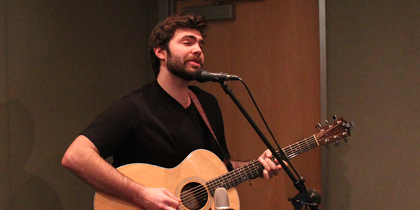 An adult playing acoustic guitar and singing into a microphone in a radio studio