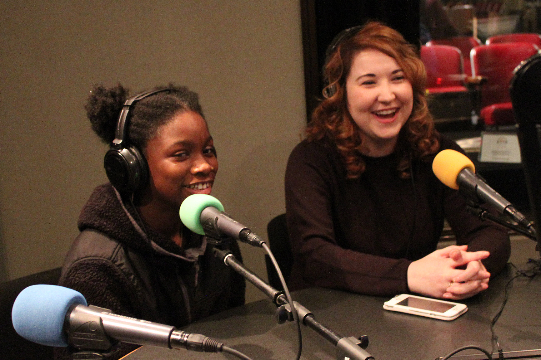 A youth and an adult wearing headphones speaking into microphones in a radio studio