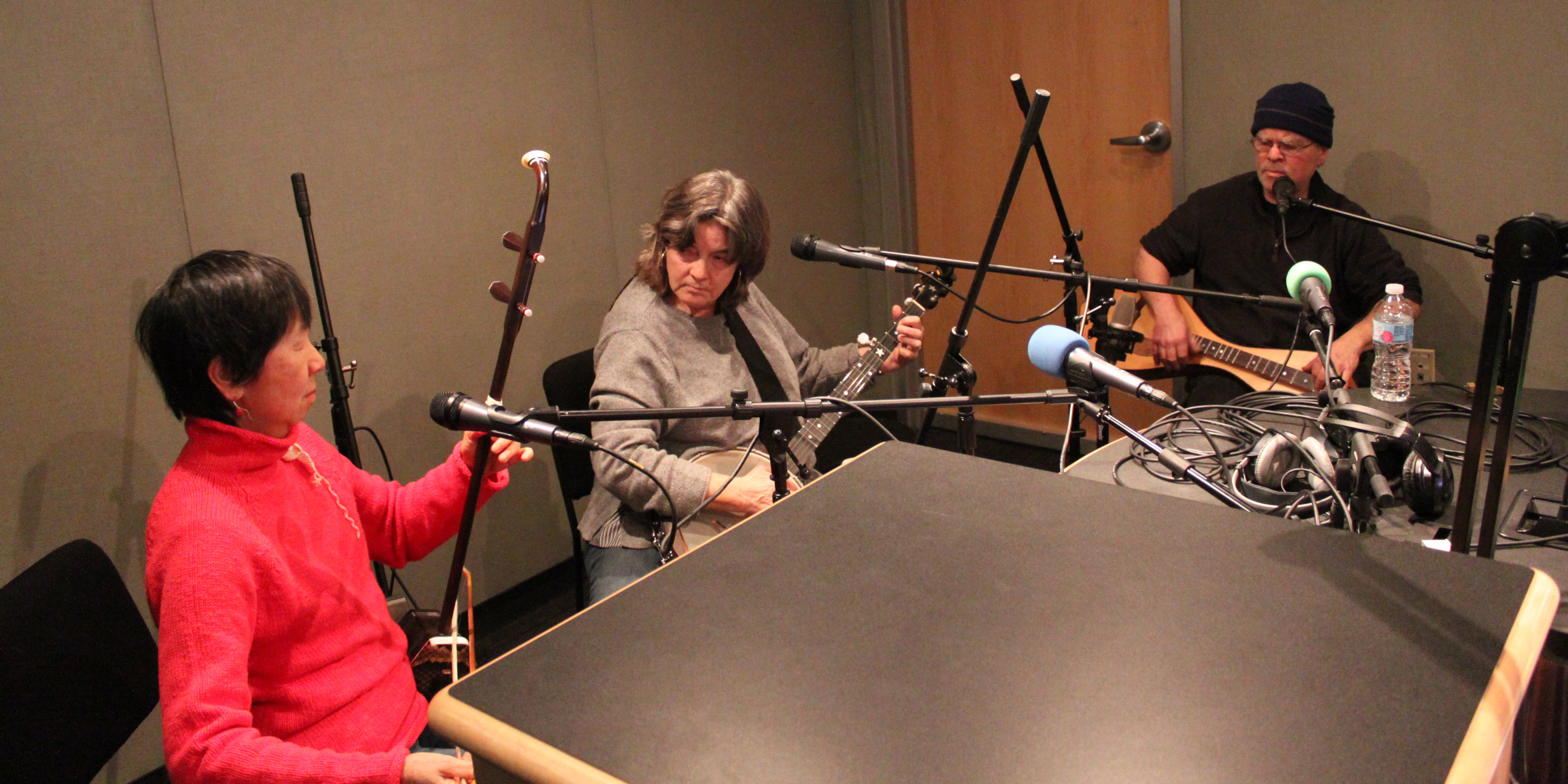 Three adults playing string instruments sitting behind microphones in a radio studio