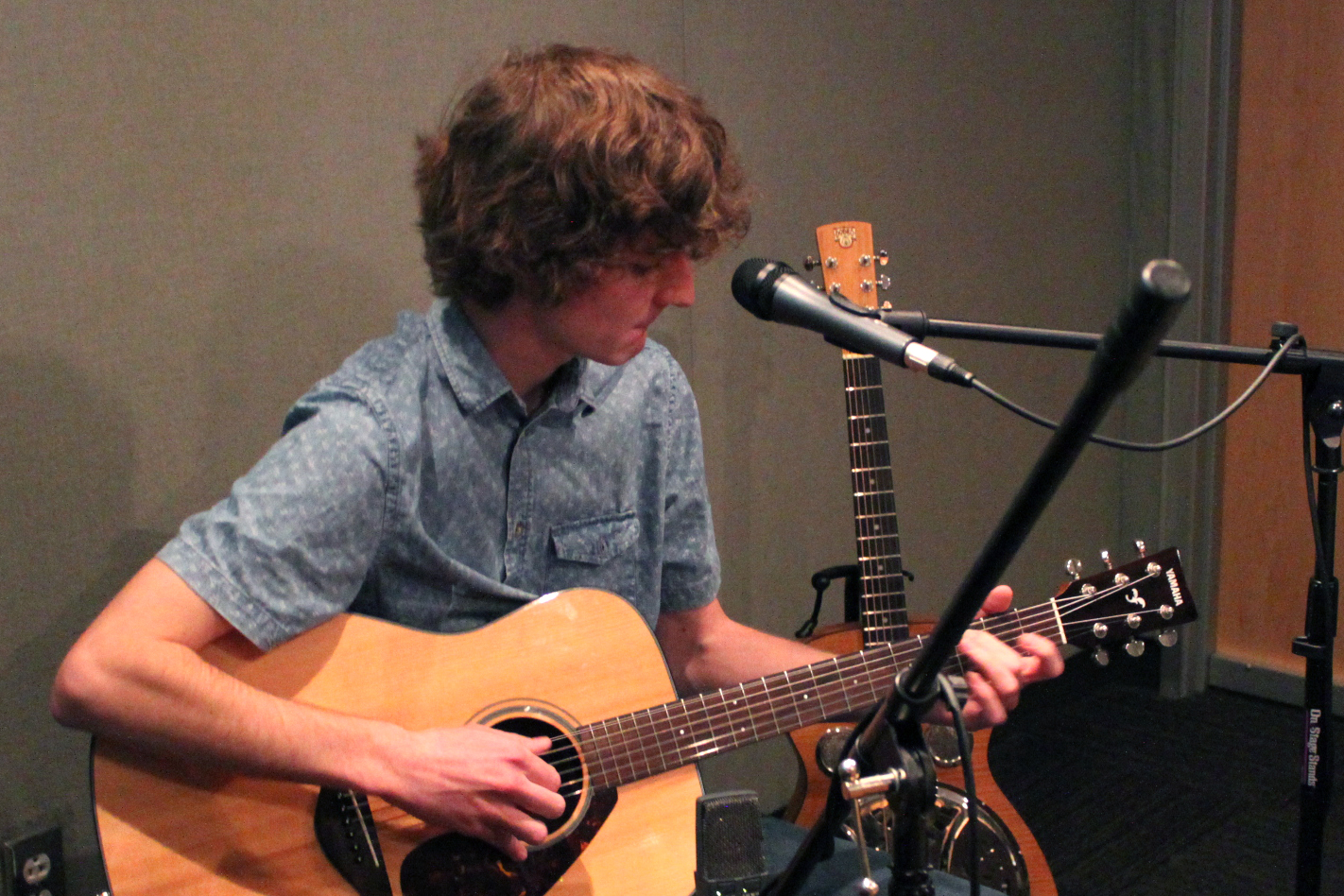 A teen playing an acoustic guitar sitting behind a microphone in a radio studio
