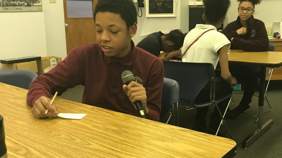 Four youth sitting around classroom desks, one holding and speaking into a microphone connected to a portable recorder