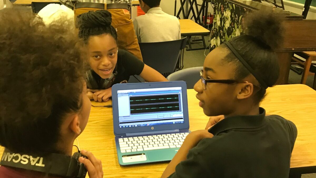 Three youth gathered around a blue laptop on a wooden classroom desk displaying waveforms in audio editing software