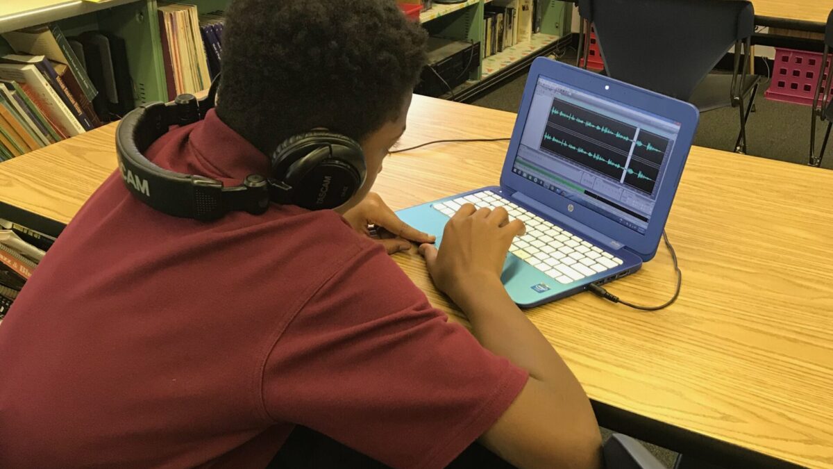 A youth wearing headphones around their neck looking at waveforms in audio editing software on a blue laptop sitting on a wooden classroom desk