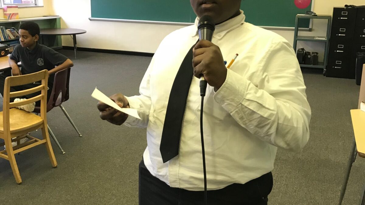 A teen holding a piece of paper and holding and speaking into a microphone in a classroom