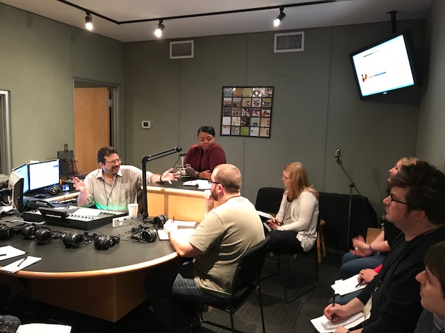 Six adults writing on pieces of paper listening to an adult gesturing with their hands sitting behind a mixing board in a radio studio