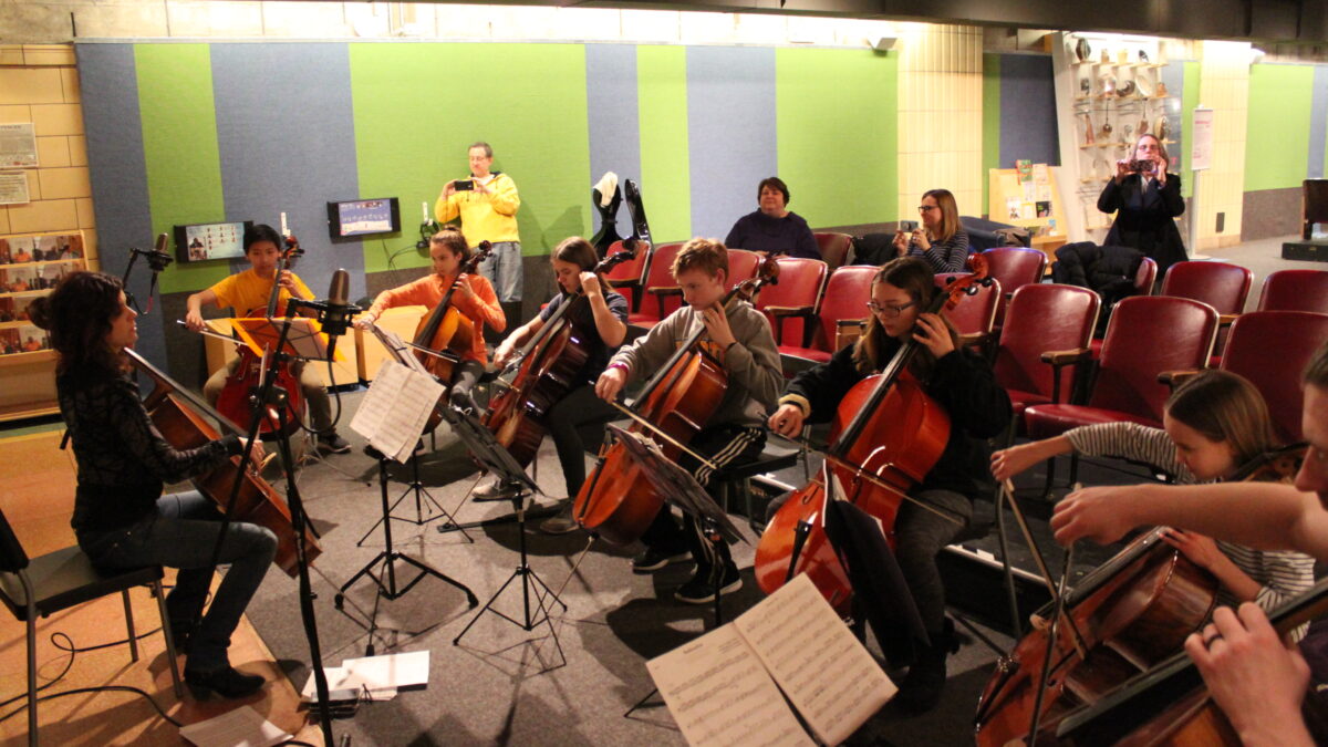 Eight individuals playing cello and looking at sheet music on music stands as four adults listen in the background, three filming on smartphones