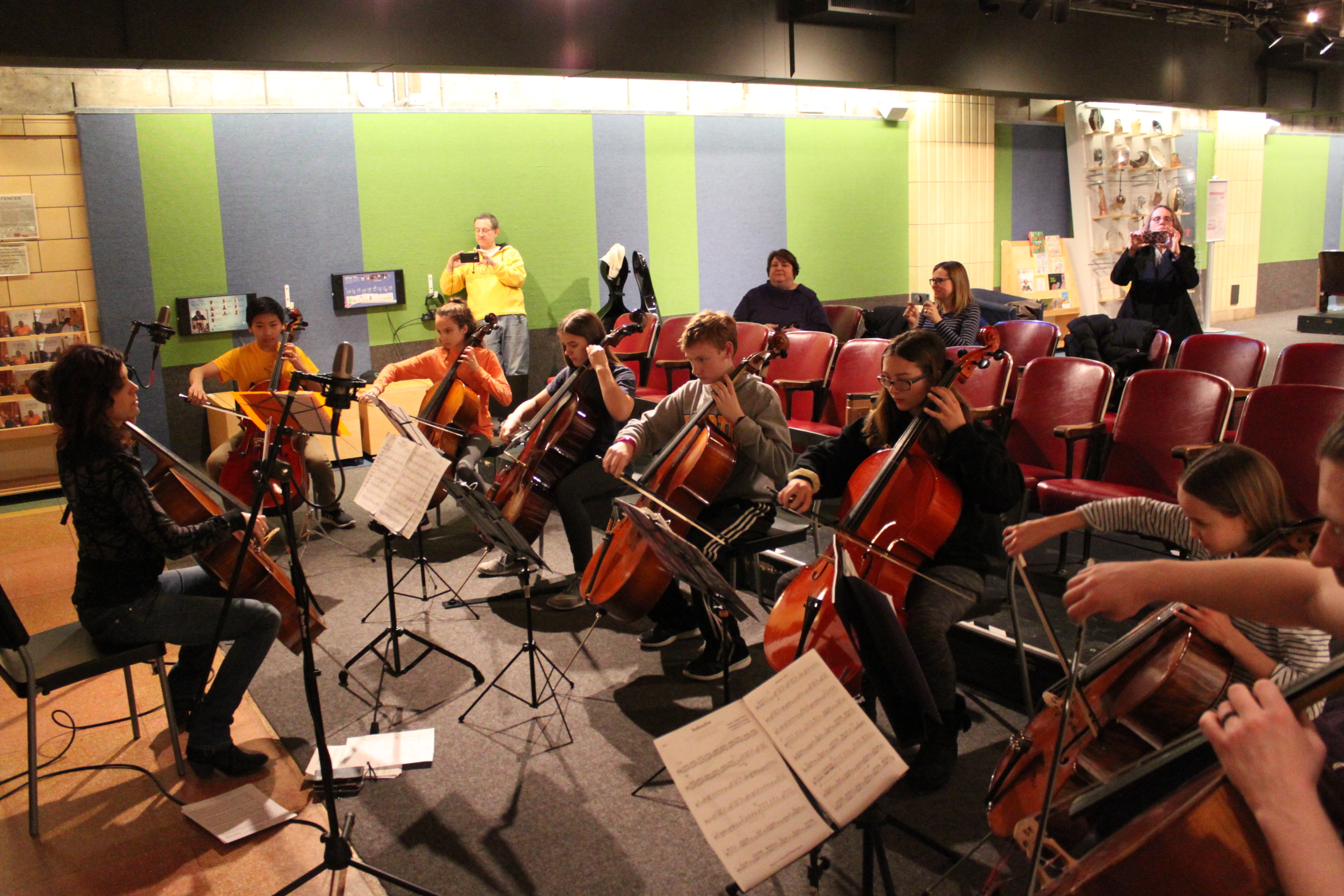 Eight individuals playing cello and looking at sheet music on music stands as four adults listen in the background, three filming on smartphones