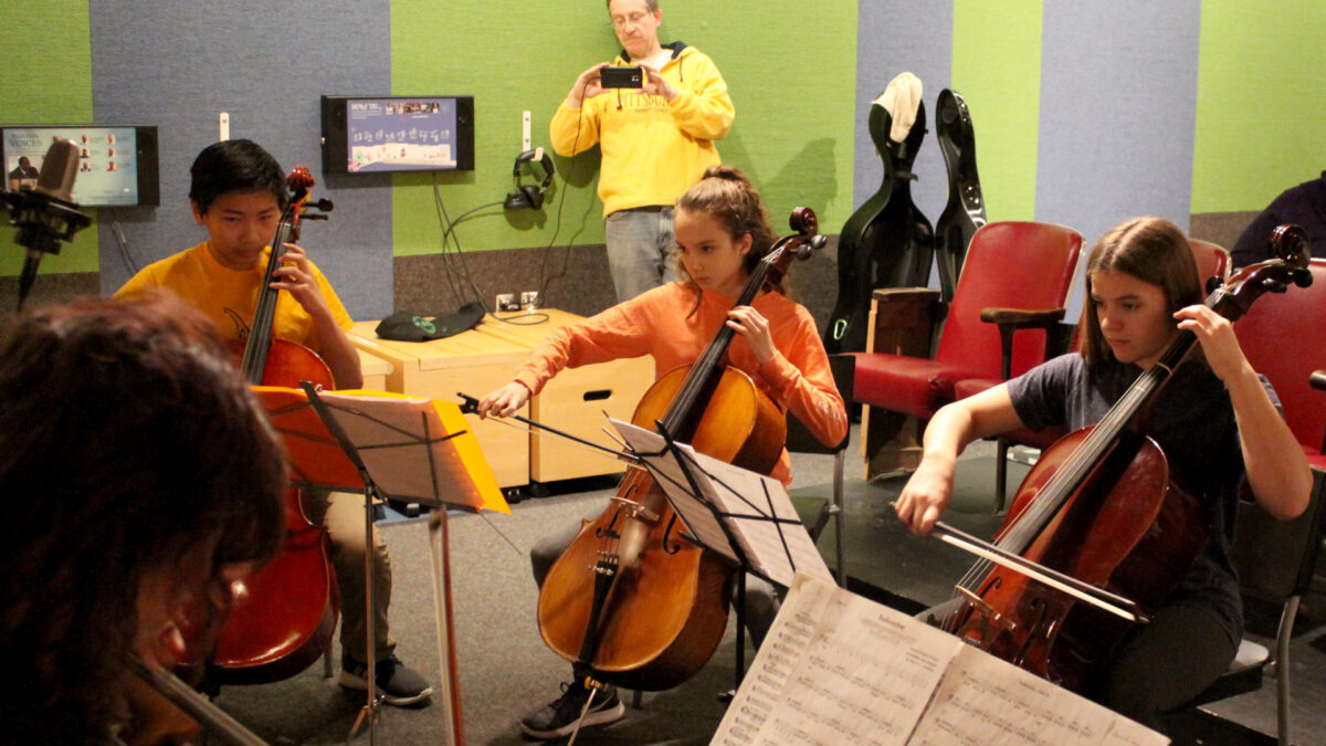 Three youth playing cello while looking at sheet music on music stands, while an adult stands in the background filming them on a smartphone